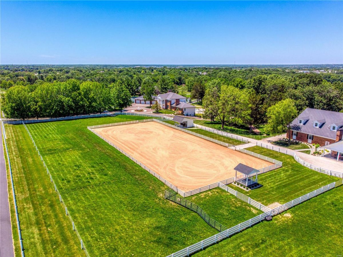 An aerial view shows a large equestrian facility. A sand riding arena is enclosed by a white fence, and grassy areas surround it.