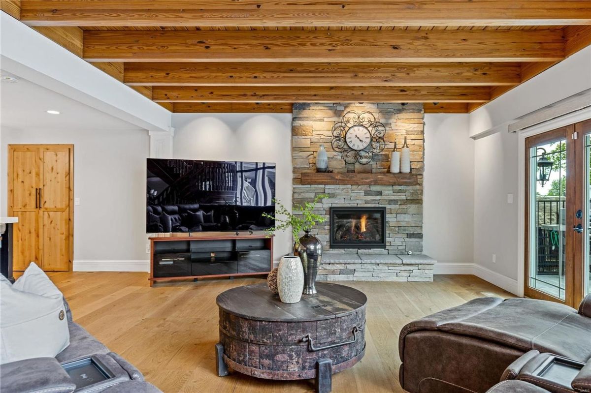 Hardwood floors and exposed wood beams highlight a living area featuring a stone fireplace, a large flat-screen television, and comfortable leather seating.