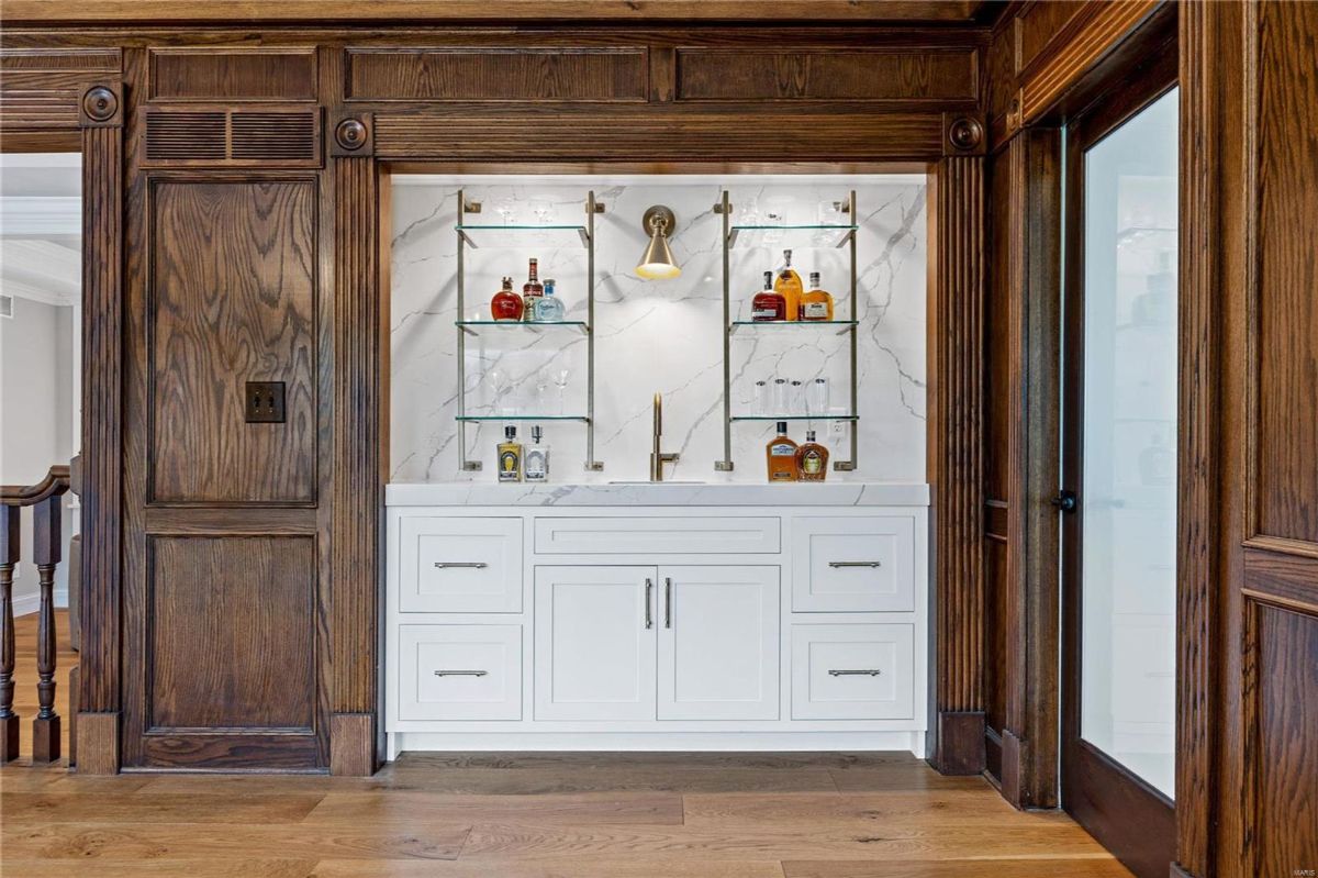 A wet bar is built into a rich, dark wood wall. It features a white cabinetry base, glass shelving, and brass accents.