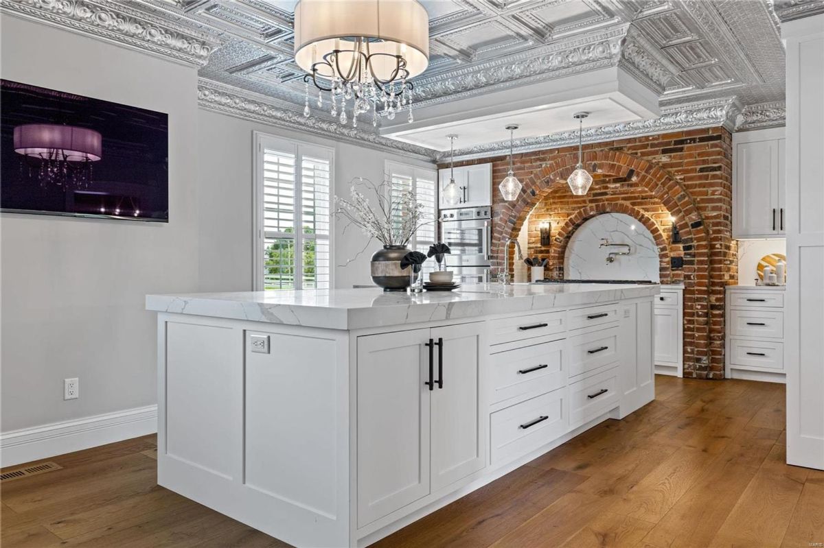 A kitchen features a large white island with a marble countertop and a brick accent wall with an arched alcove. 