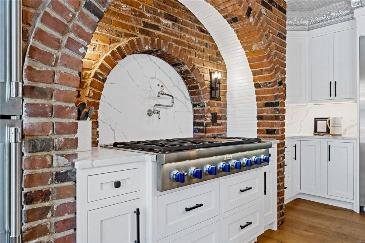 A kitchen range is set within an arched alcove of exposed brick. White cabinetry surrounds the range, which has blue knobs. 