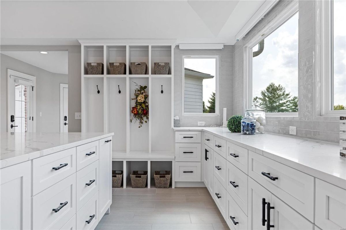 A bright, spacious laundry room or mudroom features white cabinetry with black hardware, a built-in bench with storage, and large windows.