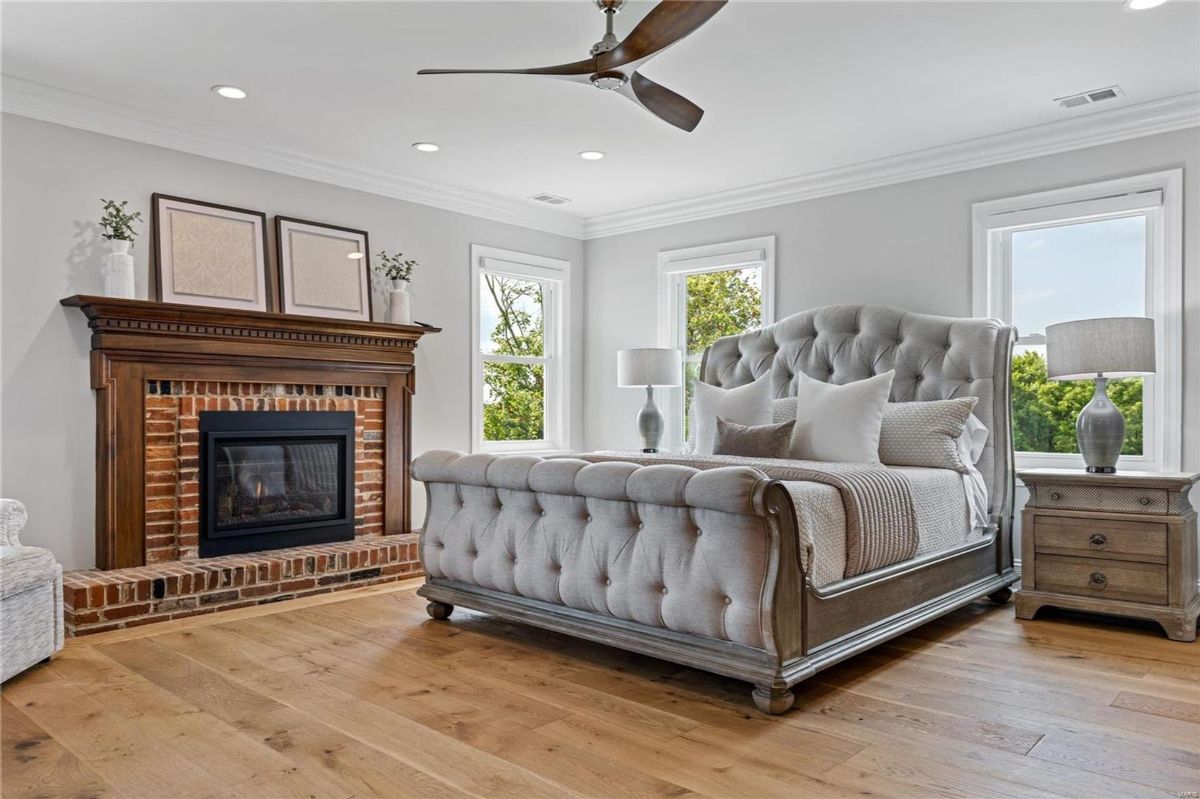 Master bedroom features a large upholstered sleigh bed, hardwood floors, and a brick fireplace with a dark wood mantel. 