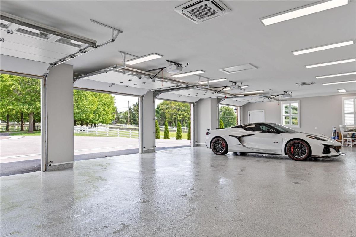 Garage with a polished concrete floor has multiple overhead doors that open to a view of trees and a driveway. 