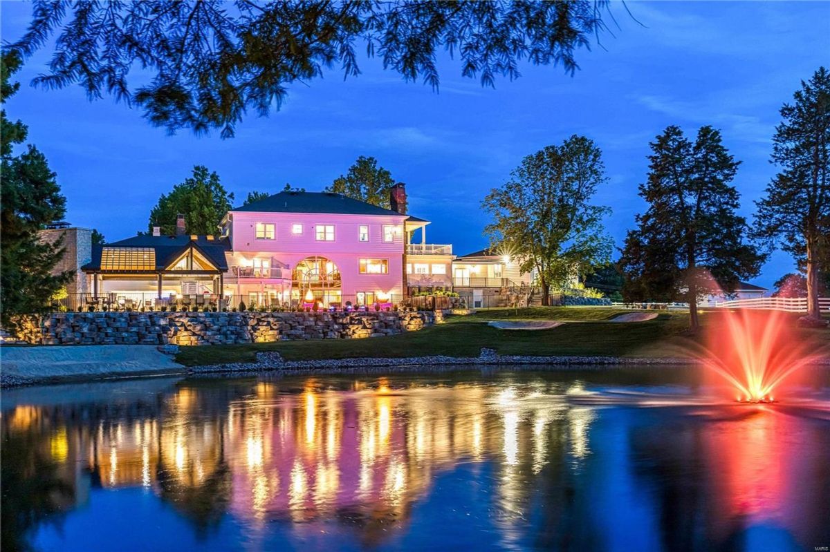 Light-pink colored home is situated on a property with a pond and fountain at night. 