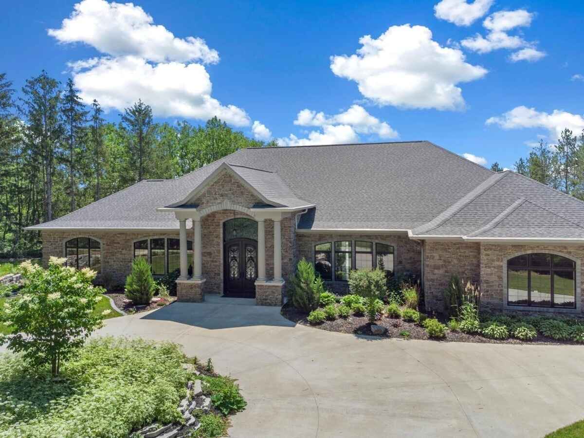 Large single-story stone house with a front porch supported by pillars, surrounded by landscaped greenery and trees.