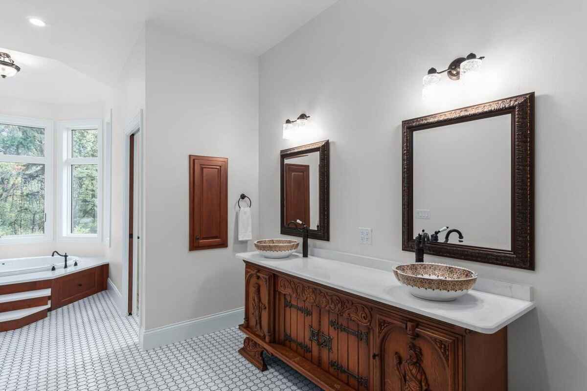 Luxurious bathroom features a double vanity with ornate wood detailing and a soaking tub surrounded by windows with a wooded view.