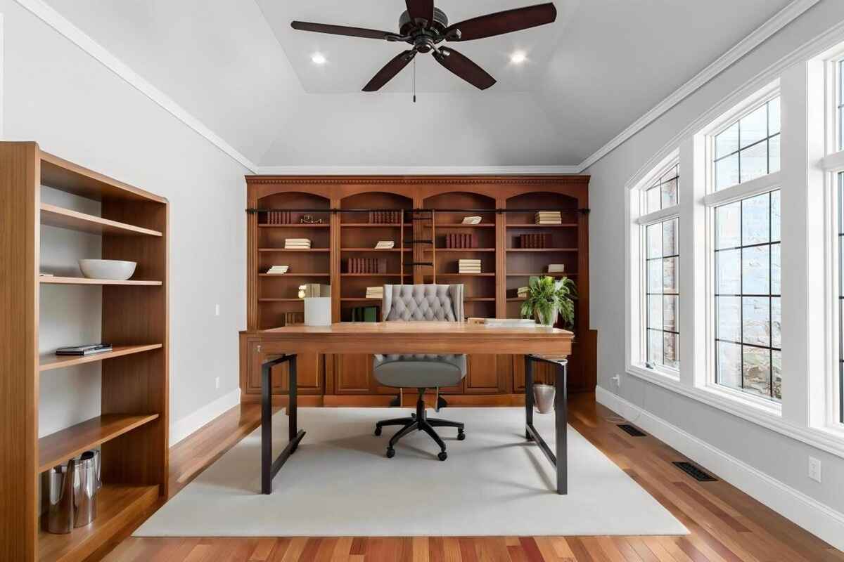 Office space contains wooden shelves, a central desk, a ceiling fan, and large windows allowing natural light.