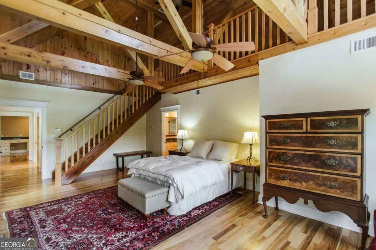 Bedroom with vaulted ceiling includes wooden beams, an exposed staircase, and a vibrant area rug.