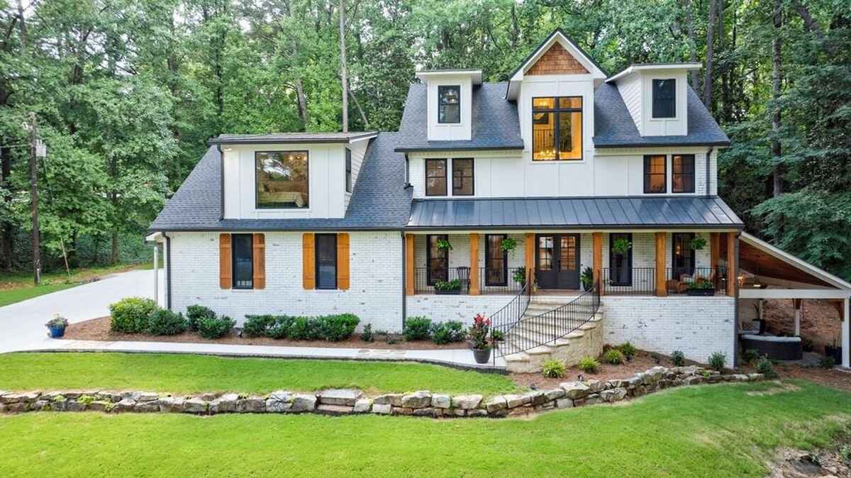  Two-story house features a white brick exterior, dormer windows, a covered porch with wooden columns, and a landscaped front yard surrounded by trees.