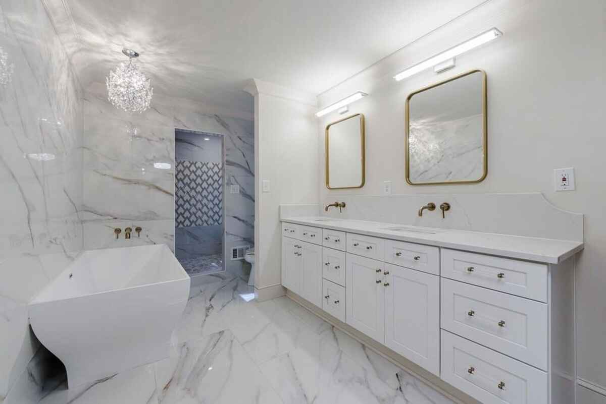 Bathroom is designed with white cabinetry, gold fixtures, marble surfaces, and a modern freestanding bathtub.