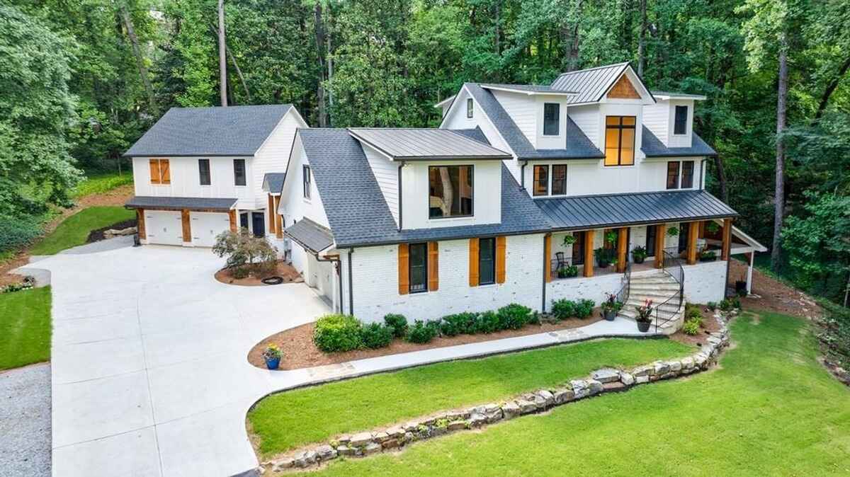  House includes a main building with a covered porch and a detached two-car garage connected by a large driveway surrounded by a wooded landscape.