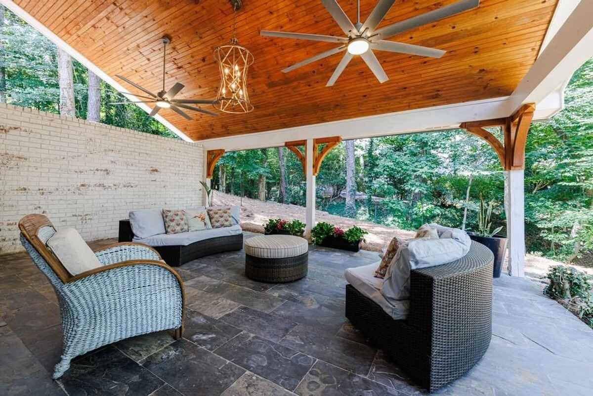 Outdoor patio furniture sits beneath a covered porch with a wood ceiling and ceiling fans, overlooking a wooded area.