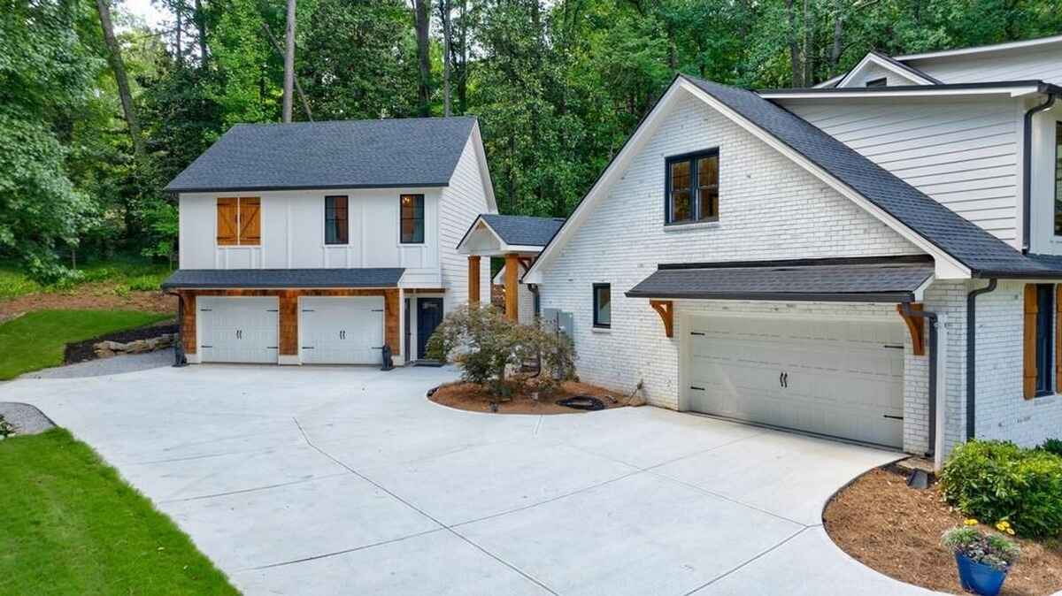 Two white houses with attached garages sit on a large concrete driveway, surrounded by lush green trees.