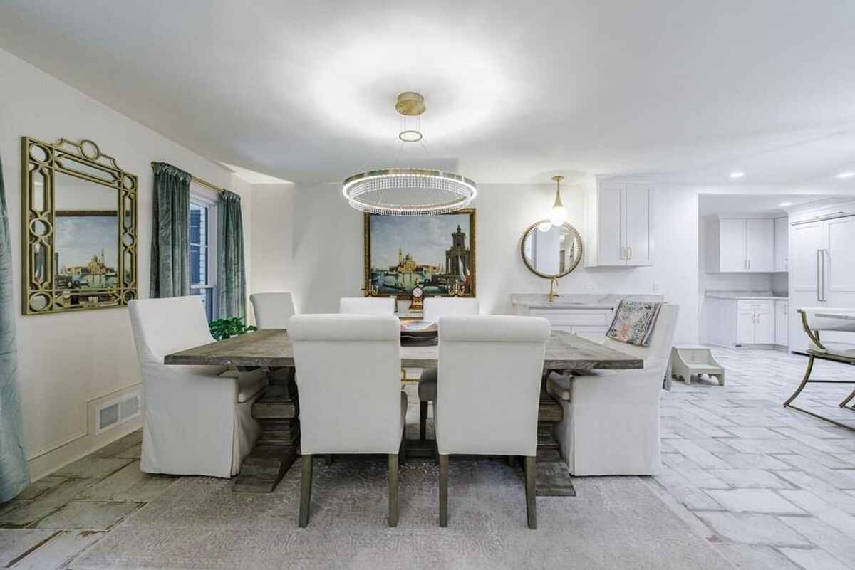 Dining area features a rectangular wooden table surrounded by white upholstered chairs, with a chandelier and framed artwork as focal points.