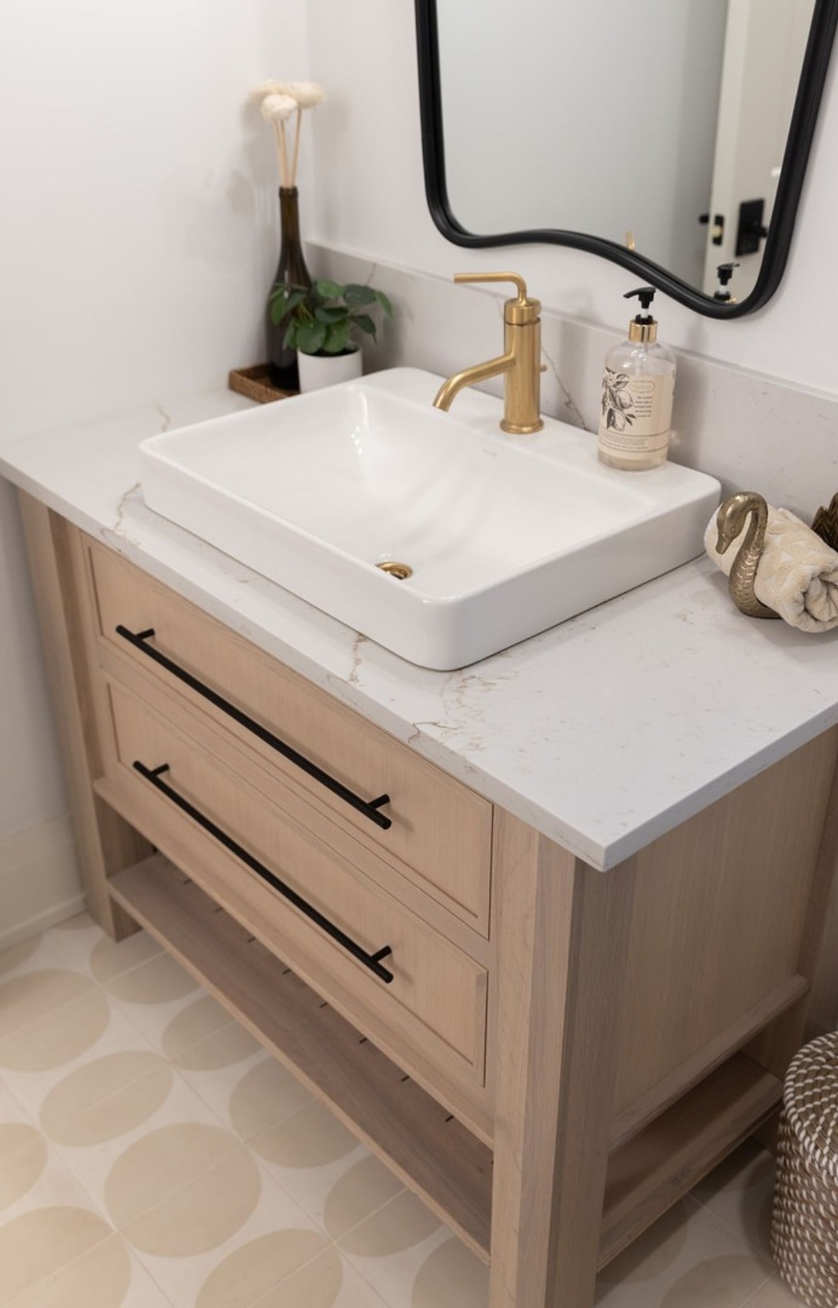 This minimalist bathroom vanity boasts a light wood finish and sleek black hardware, creating a modern and airy feel. The unique vessel sink and gold faucet add a touch of sophistication and visual interest.