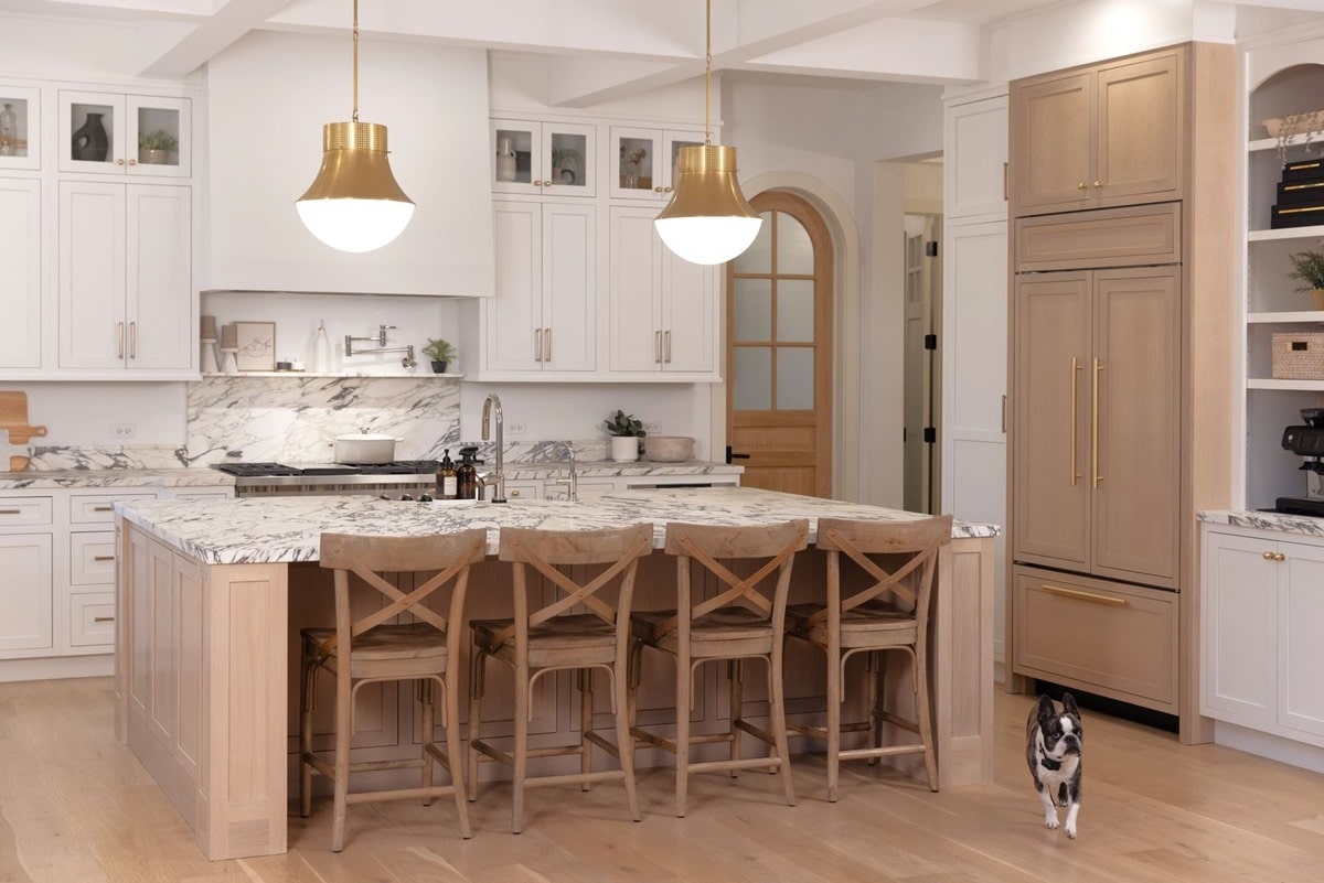 This kitchen embodies a sophisticated blend of farmhouse and transitional styles, showcasing a light and airy aesthetic. The natural wood tones of the cabinetry and island stools are beautifully complemented by the marble countertops and brass hardware.