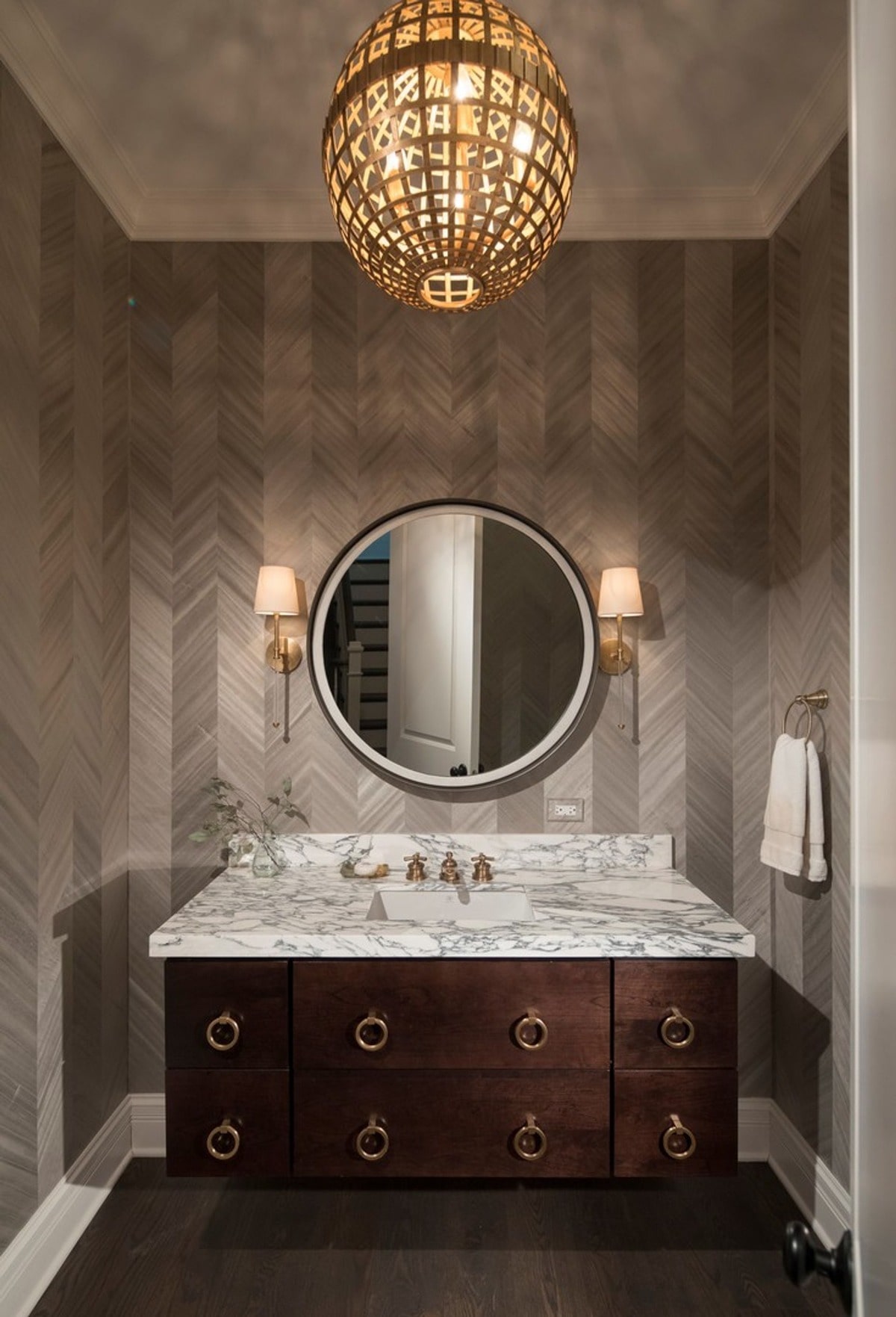 he powder room's design showcases meticulous attention to detail, from the herringbone wallpaper to the ornate light fixture and brass hardware. The combination of textures and materials creates a luxurious and inviting space.