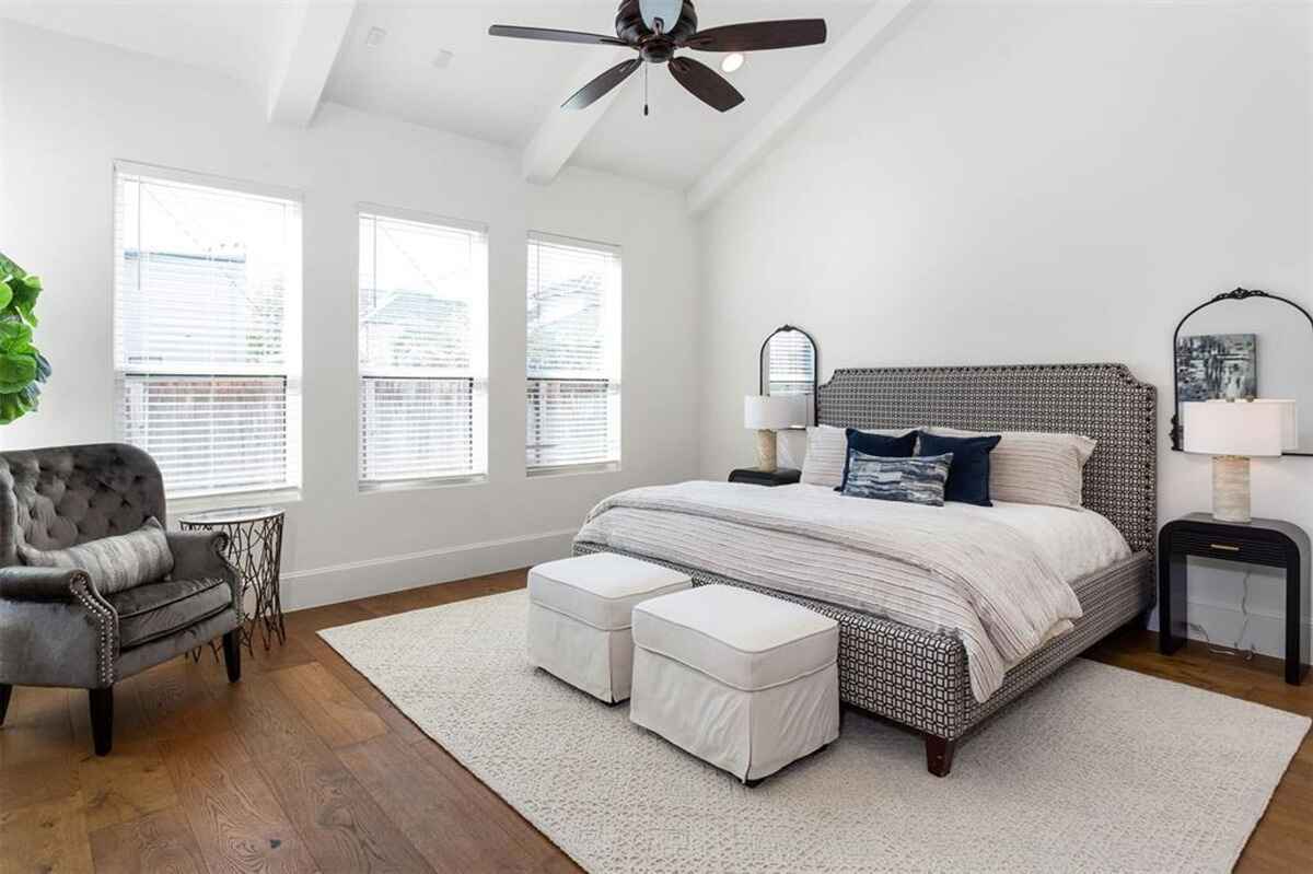 Bedroom features a patterned headboard, white ottomans, hardwood flooring, and large windows with blinds.