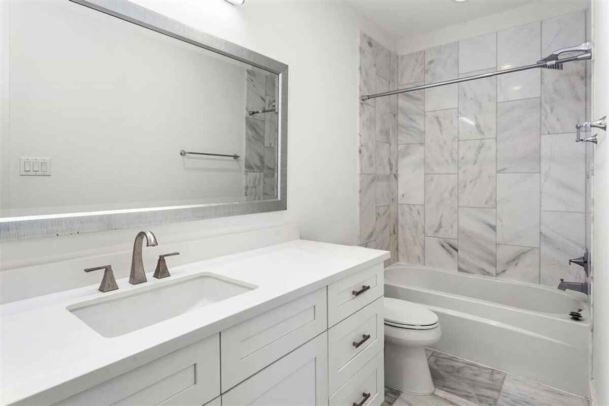 Bathroom features a white vanity with a rectangular sink, a large mirror with a metallic frame, and a bathtub with marble tile surround.