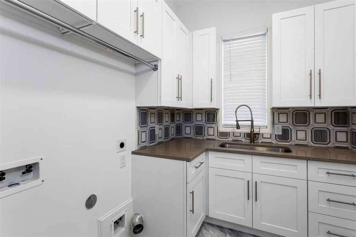 Laundry room features white cabinetry, a patterned backsplash, a stainless steel sink, and connections for a washer and dryer.