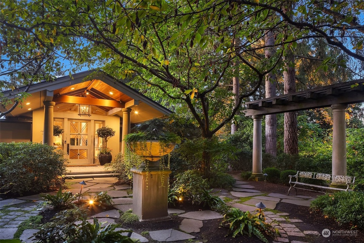 A beautifully lit house entrance is shown, featuring a stone pathway, a pergola, and a bench, all set within a lush, tree-filled landscape.