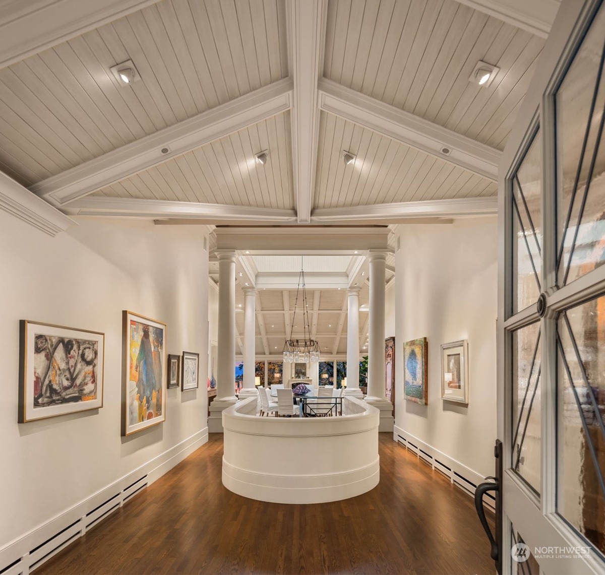 A long hallway with white walls and a white balustrade displays artwork, leading to a spacious room with a high, beamed ceiling and a chandelier.