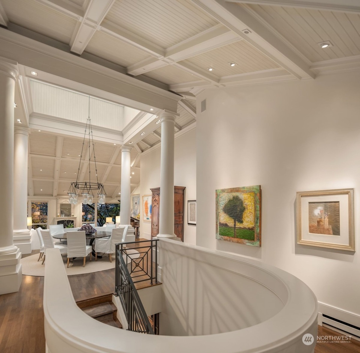 A curved hallway with white walls and columns overlooks a formal dining area below, showcasing artwork and a high ceiling with a skylight.