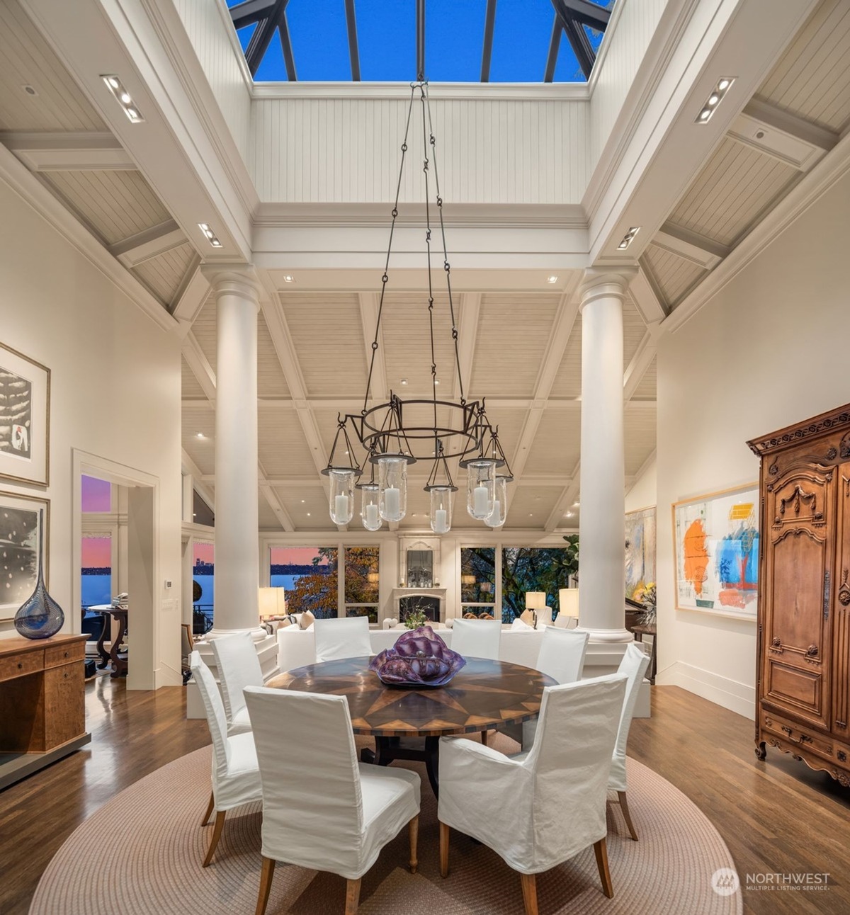 A formal dining room with a large round table, white chairs, and a dramatic chandelier is highlighted by a skylight and tall columns.