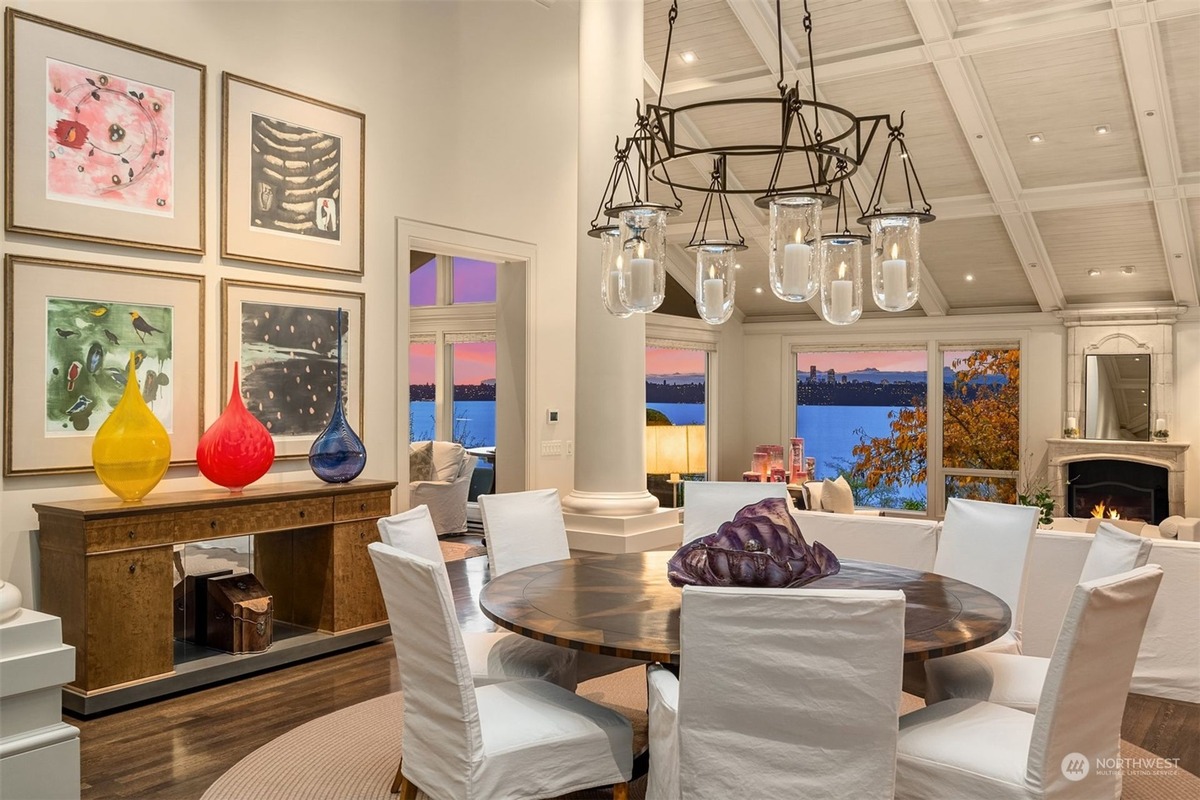 An elegant dining room features a large round table surrounded by white slipcovered chairs, a stunning water view from large windows, and artwork adorning the walls.