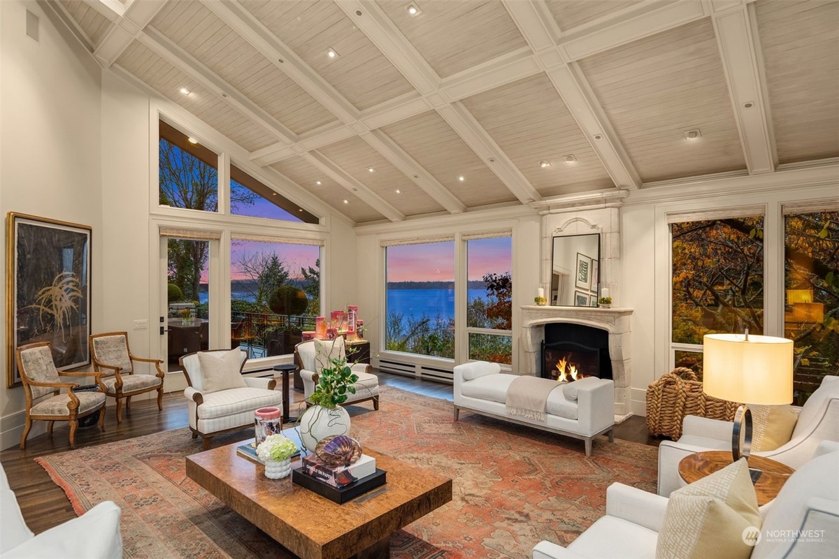 A luxurious living room features a high ceiling with exposed beams, expansive windows showcasing a captivating water view, and tastefully arranged furniture.