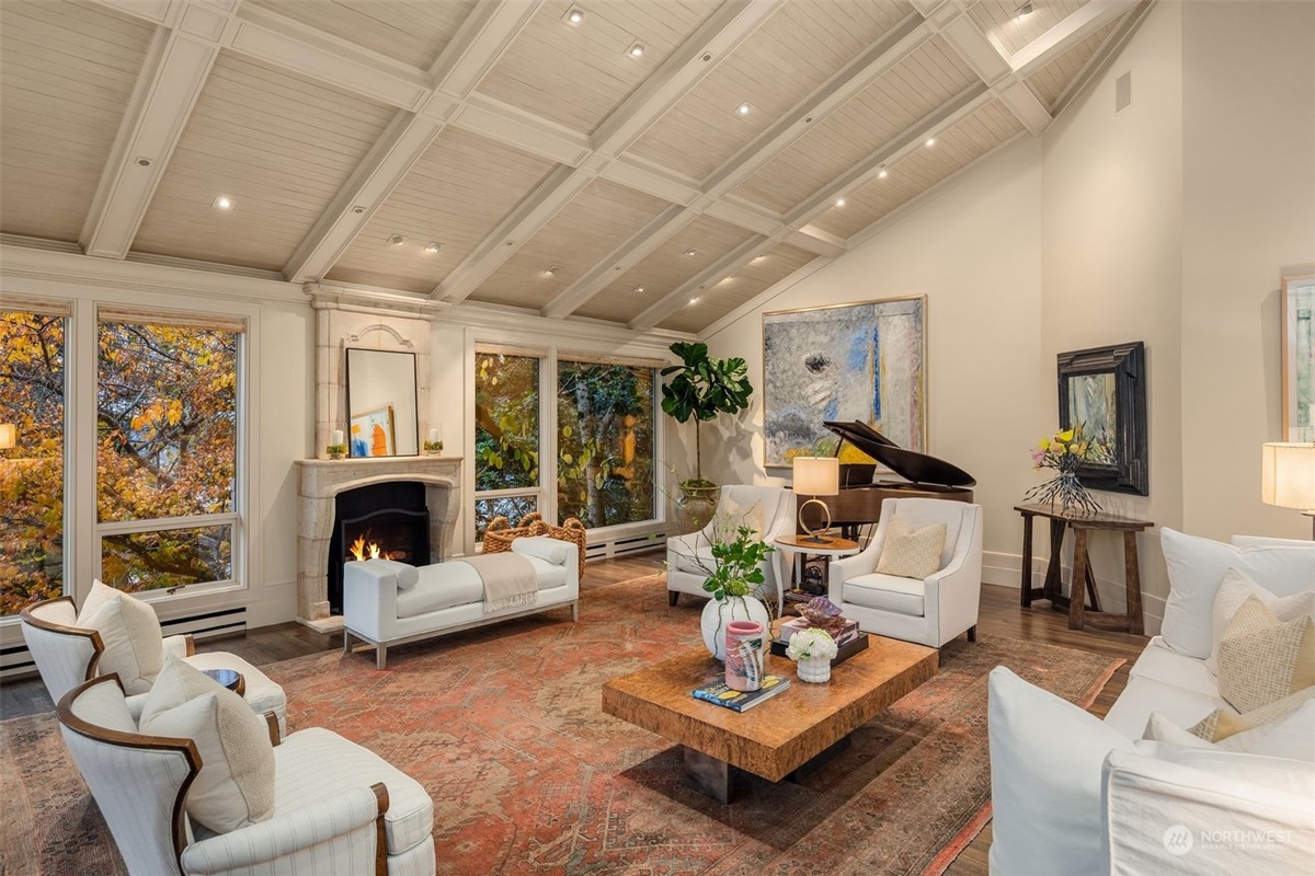 A high-ceilinged living room, featuring a fireplace and large windows overlooking autumnal trees, is elegantly furnished with white sofas, armchairs, and a grand piano.