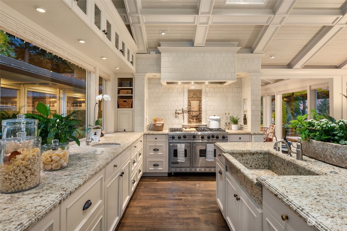 A bright, spacious kitchen with white cabinetry and granite countertops features a large island with a farmhouse sink, a professional-style range, and a view of the outdoors through large windows.