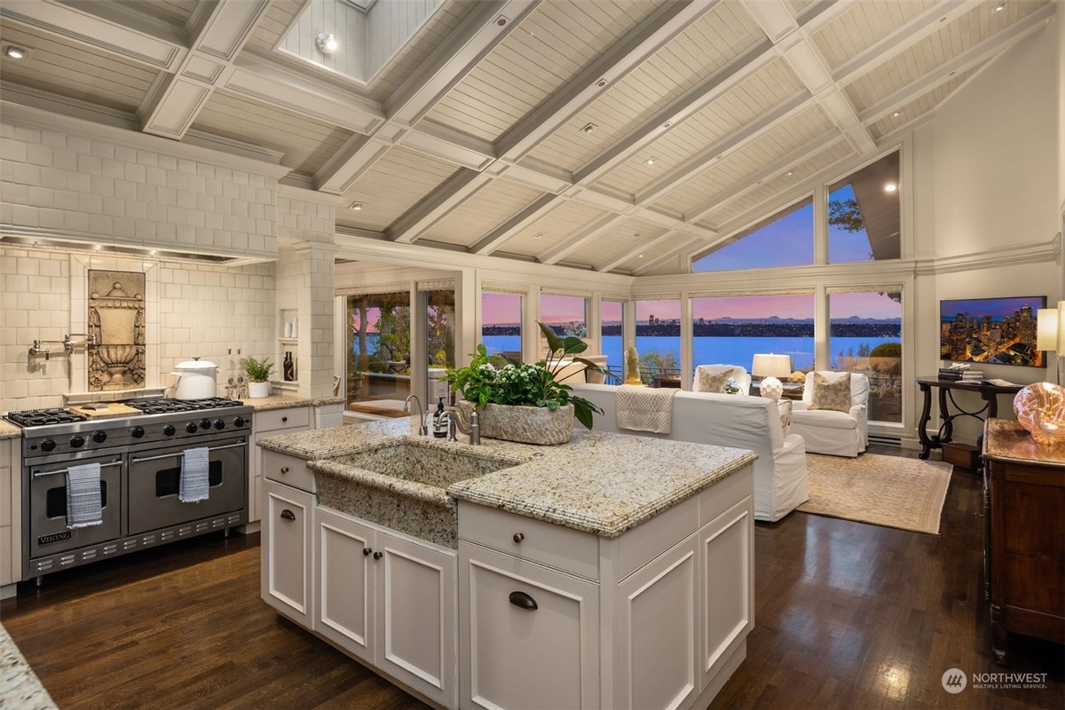 A bright kitchen with white cabinets and granite countertops is shown, featuring a large island and a professional-grade range, all overlooking a stunning waterfront view through expansive windows.