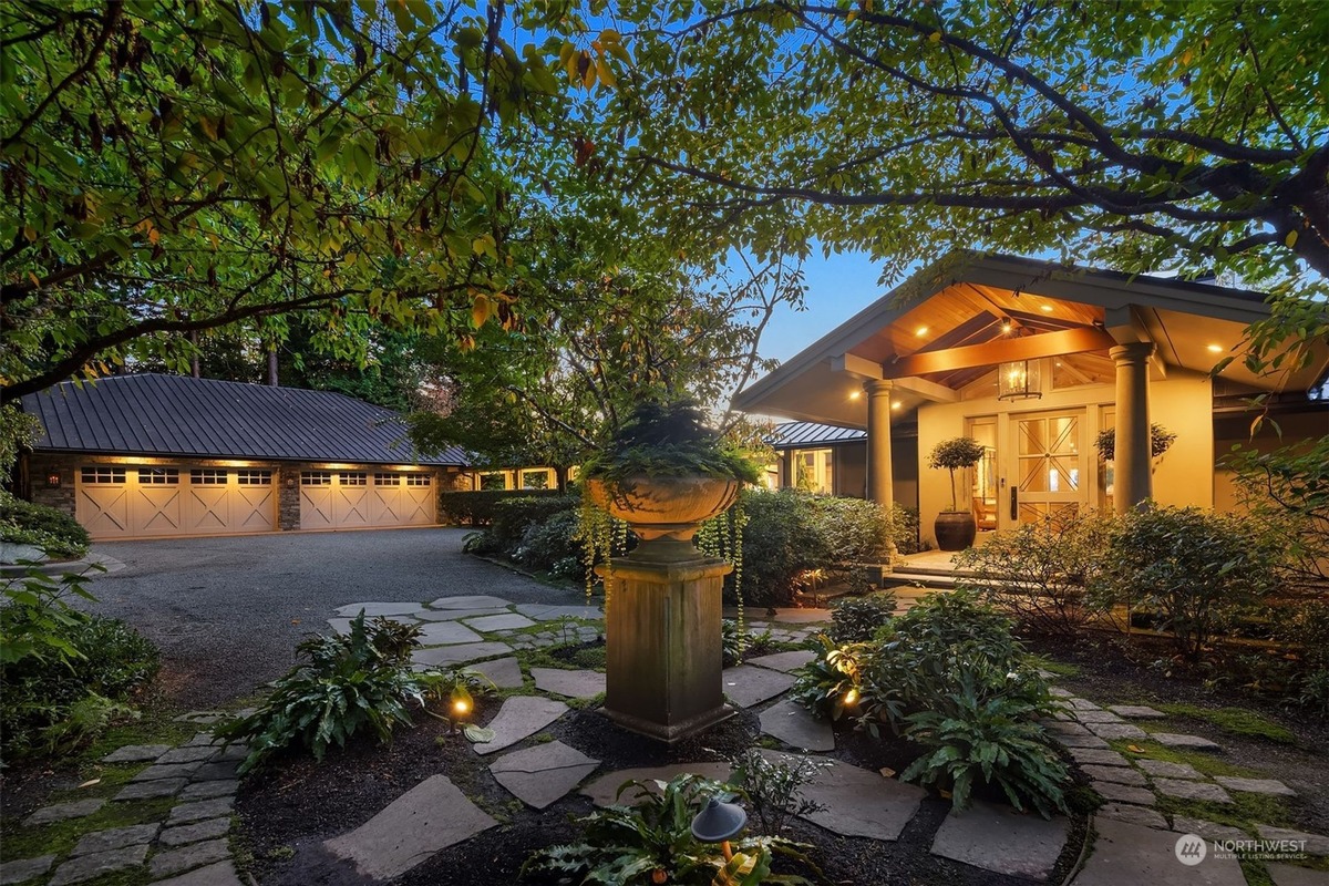 A grand house with a three-car garage is showcased, its entrance adorned with a stone pathway and a decorative urn, all nestled amidst lush greenery.