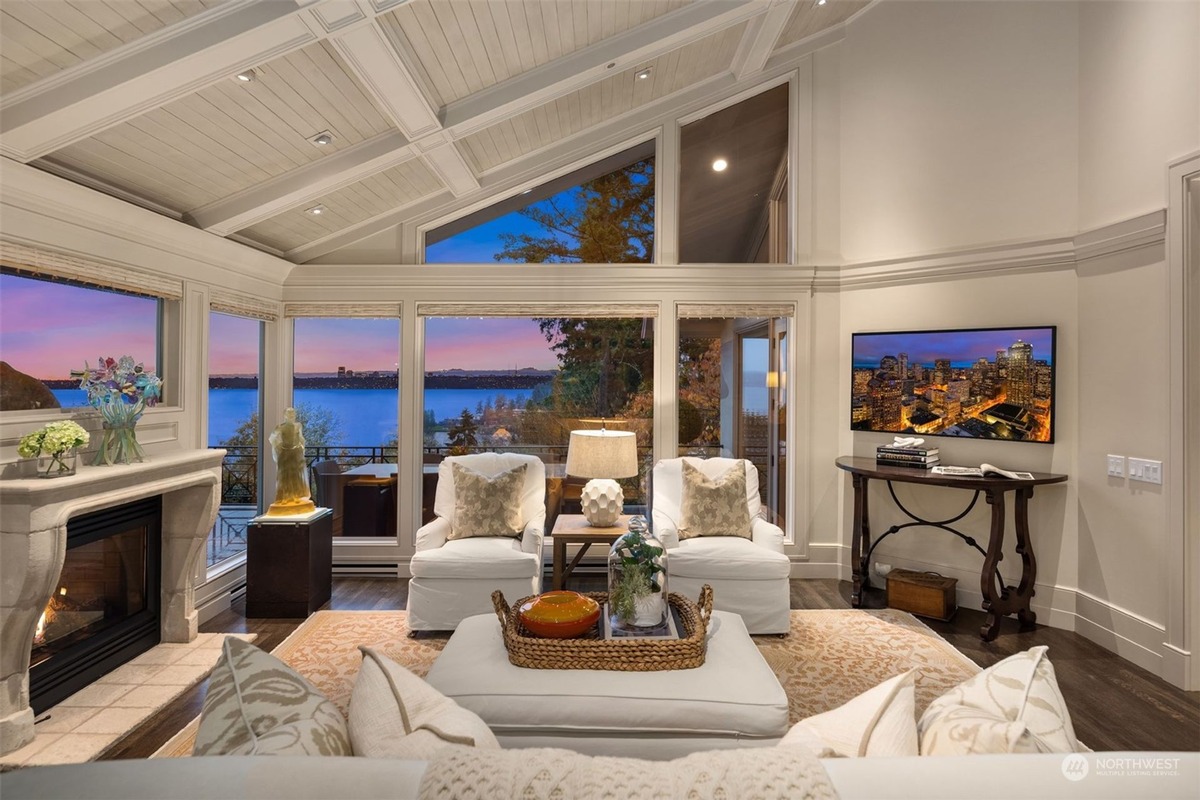 A living room with a vaulted ceiling and expansive windows offering a breathtaking sunset view over a body of water is furnished with comfortable white sofas and armchairs.