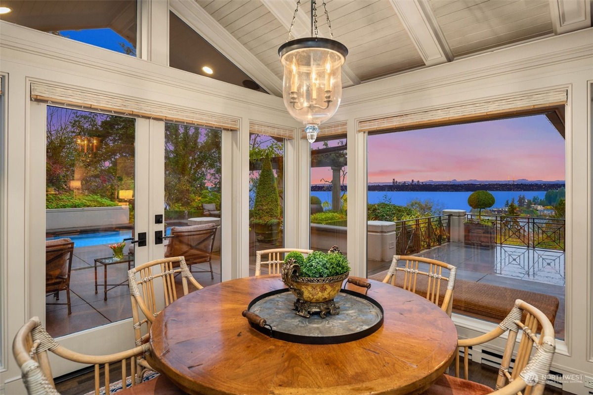 A dining room with a large wooden table and chairs offers a stunning view of a pool and waterfront scenery through expansive windows.