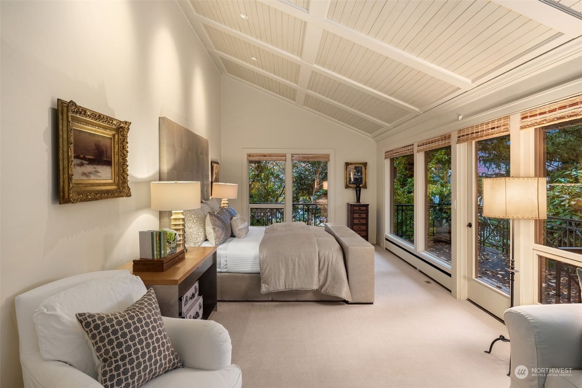 A master bedroom with a high, beamed ceiling and large windows that offer a view of trees and water features a bed with light-colored bedding and an armchair with a patterned pillow.