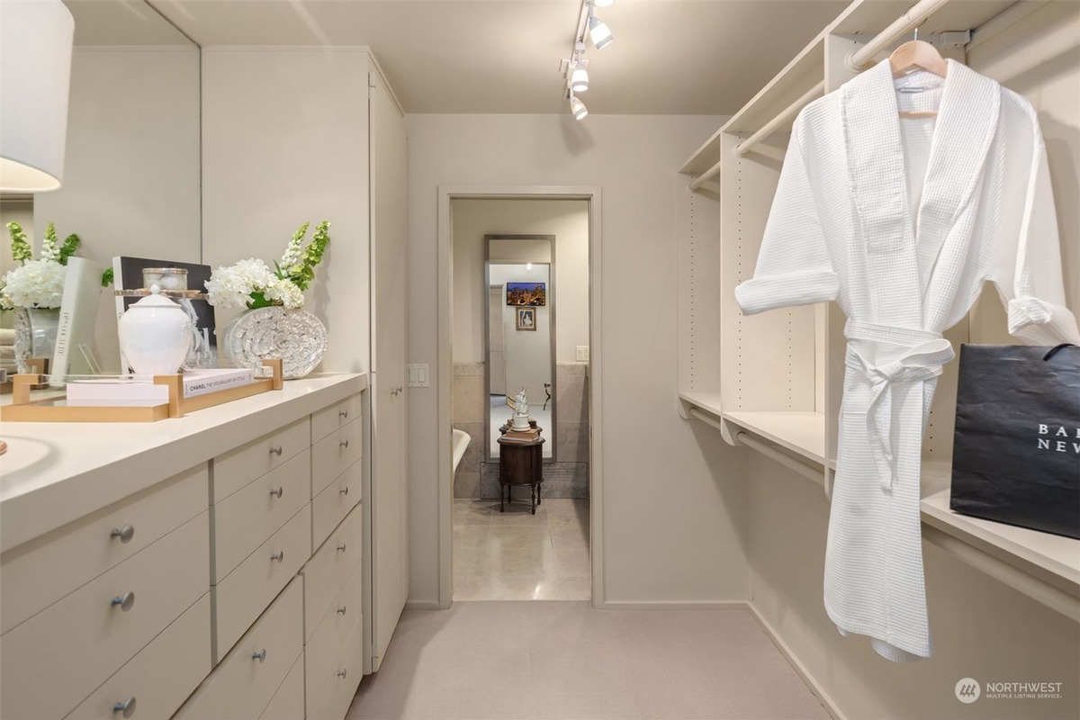 A spacious walk-in closet with light-colored built-in cabinetry and a white bathrobe hanging neatly is shown, featuring a dressing table with flowers and a view of a bathroom beyond.