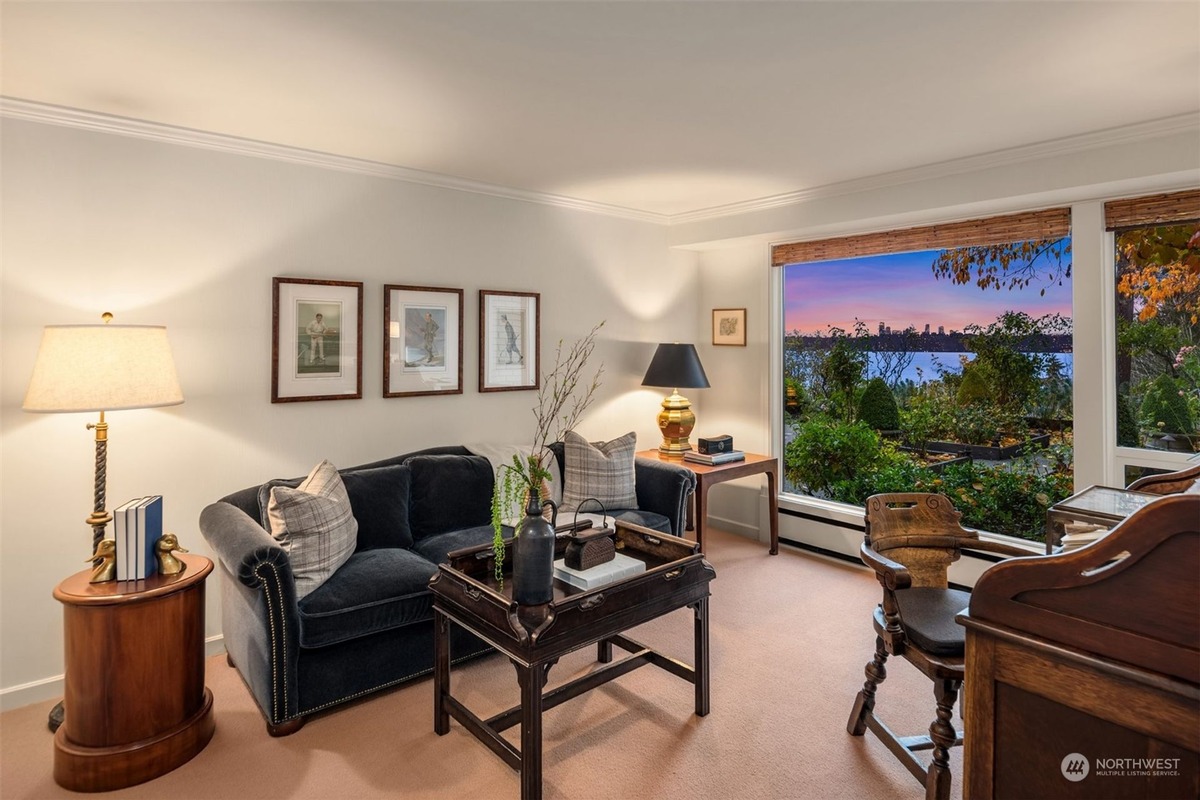 A living room with a navy blue sofa and a wooden coffee table is shown, featuring framed artwork on the walls and a large window offering a scenic view of water and cityscape at sunset. A wooden desk and chair are also visible.