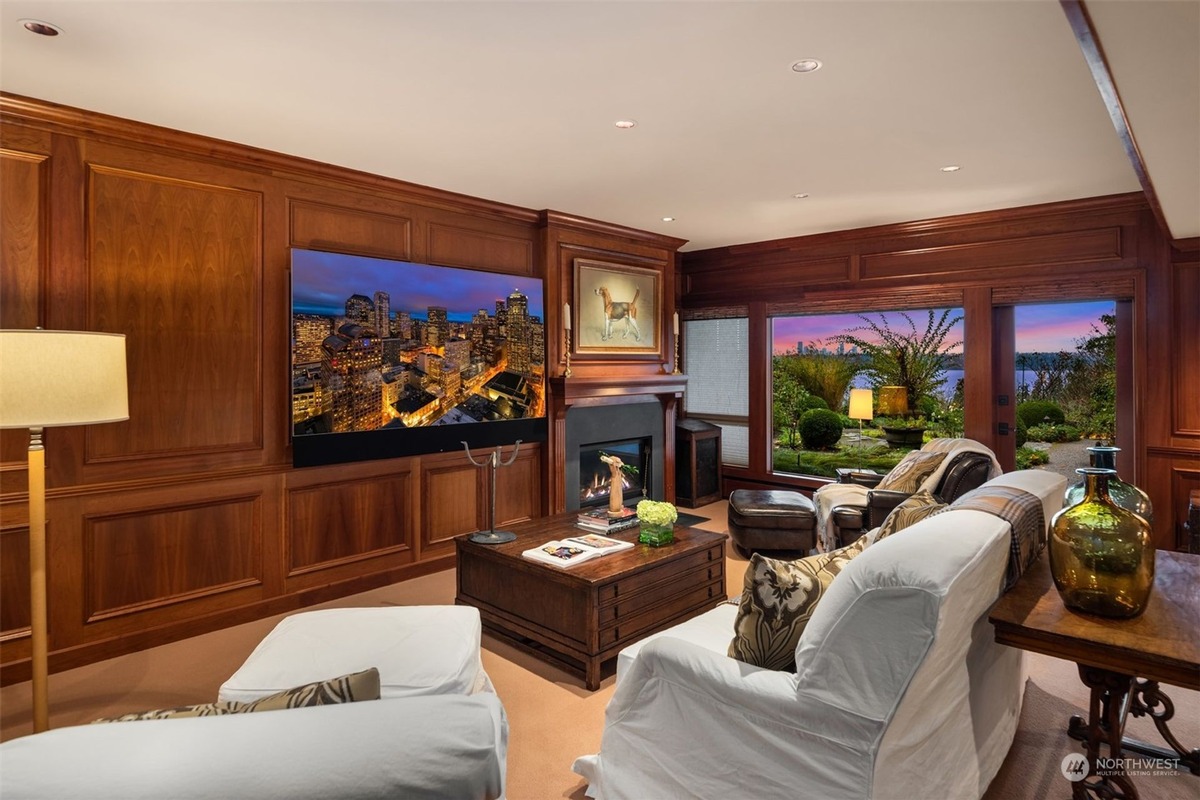 A family room with wood-paneled walls and a large-screen television is shown, featuring a comfortable seating area with a view of a garden and water beyond large windows.