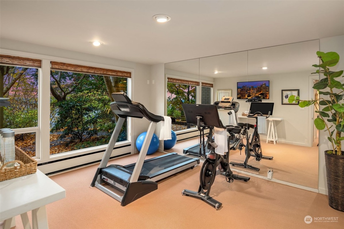 A home gym with a treadmill, exercise bike, and weights is shown, featuring large windows offering a view of trees and water. A mirror reflects the room.