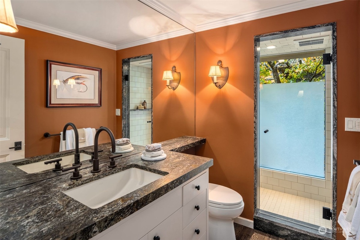 A bathroom with orange walls and a dark granite countertop vanity features a double sink with black faucets and a separate shower stall with a frosted glass door. Artwork hangs on the wall.