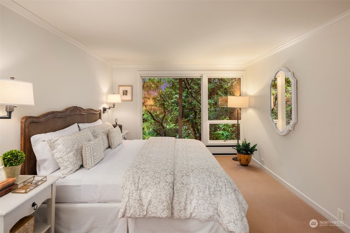 A bedroom with a wooden headboard bed dressed in white linens is shown, featuring a large window overlooking lush greenery and a decorative mirror on the wall.