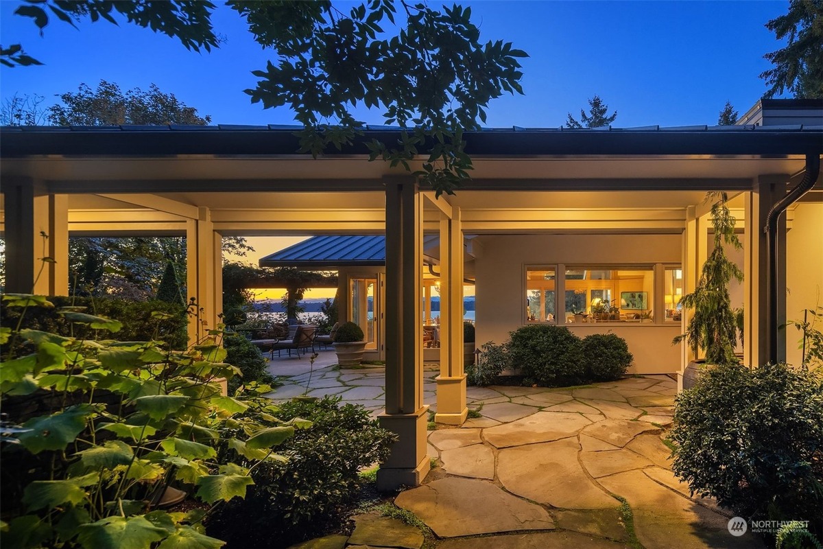 A flagstone patio area with landscaping and lighting is shown at dusk, offering a view of a house with large windows and a water view beyond.