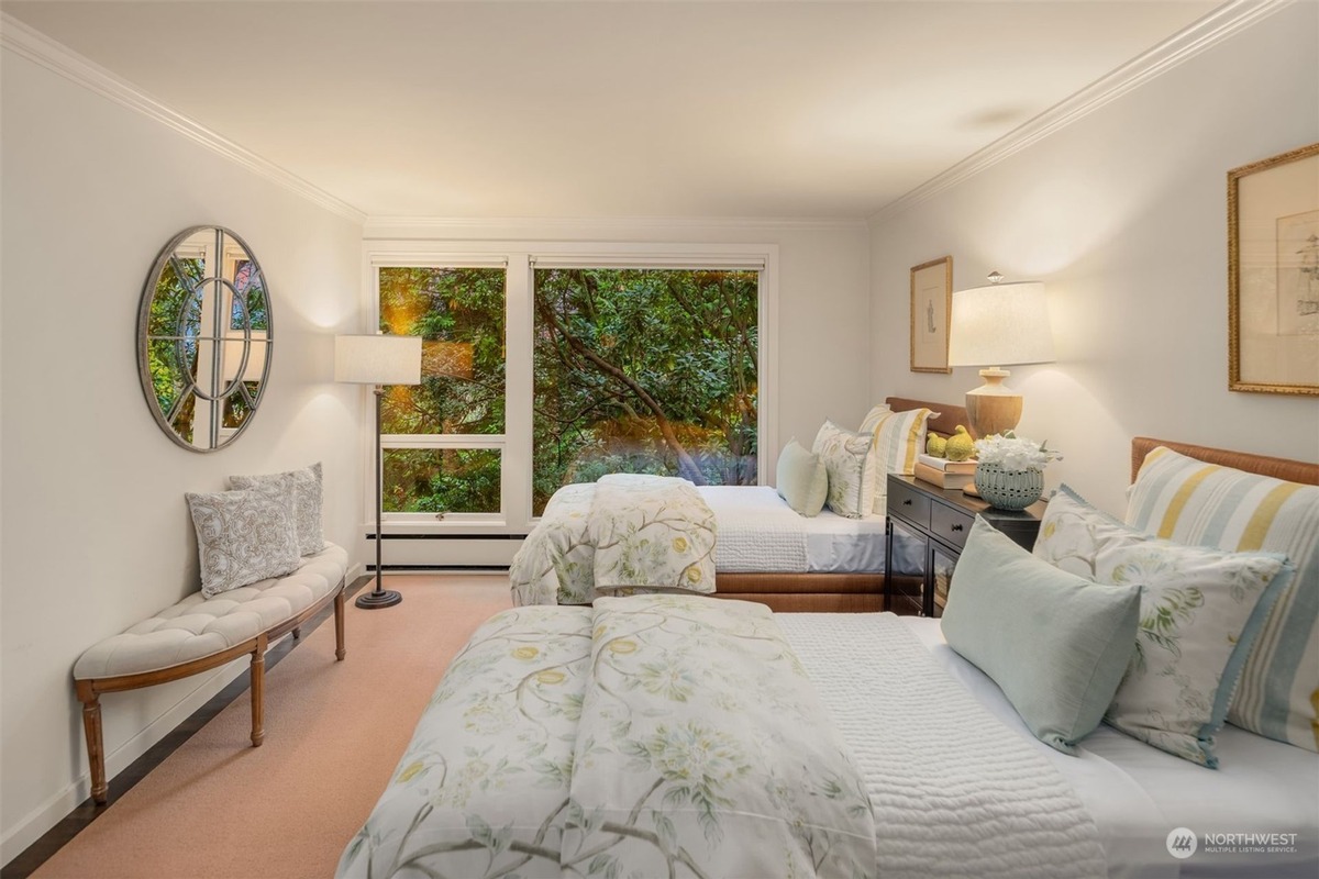 A bedroom with two twin beds dressed in light-colored bedding features a large window overlooking trees, a curved bench, and artwork on the walls.