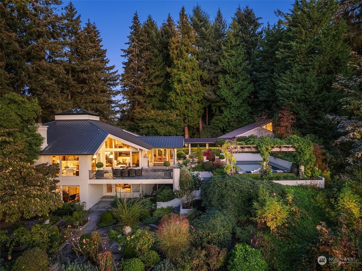 An aerial view shows a luxurious white house nestled among lush greenery and towering evergreen trees, complete with a swimming pool and meticulously landscaped gardens.