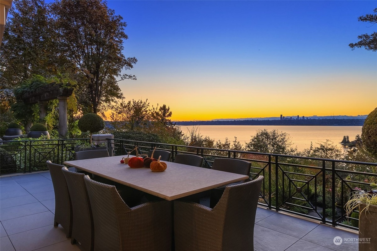 An outdoor patio dining area with a large table and chairs offers a breathtaking sunset view over a body of water and a cityscape in the distance. Ornamental plants and a railing surround the patio.