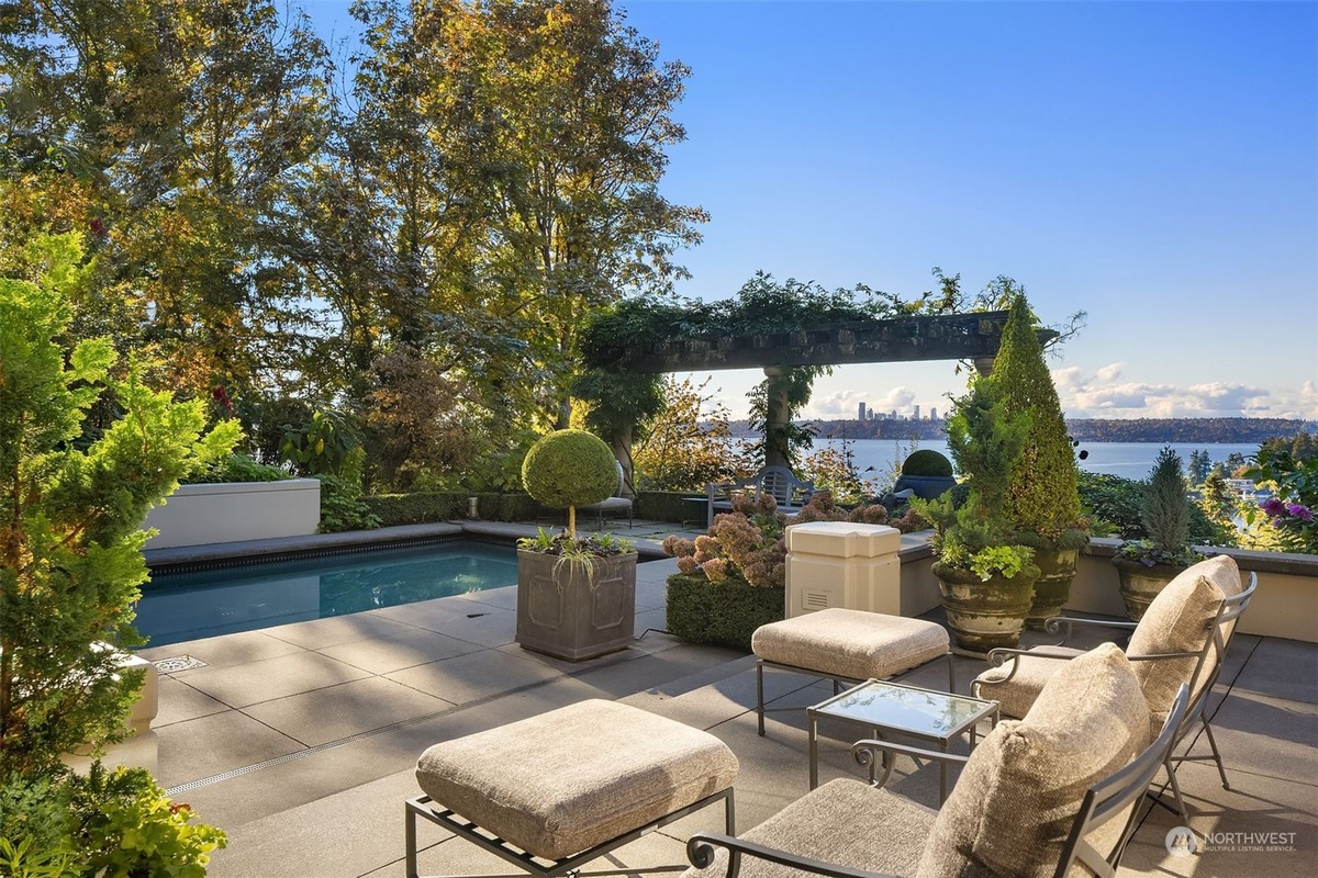 A backyard patio with lounge chairs and a swimming pool offers a stunning view of a body of water and a distant cityscape under a clear sky. Landscaping and a pergola add to the scene.