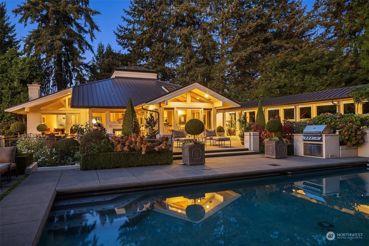 A house with a swimming pool and outdoor kitchen is shown at night, surrounded by landscaping and trees. The house is illuminated, and its reflection is visible in the pool's water.